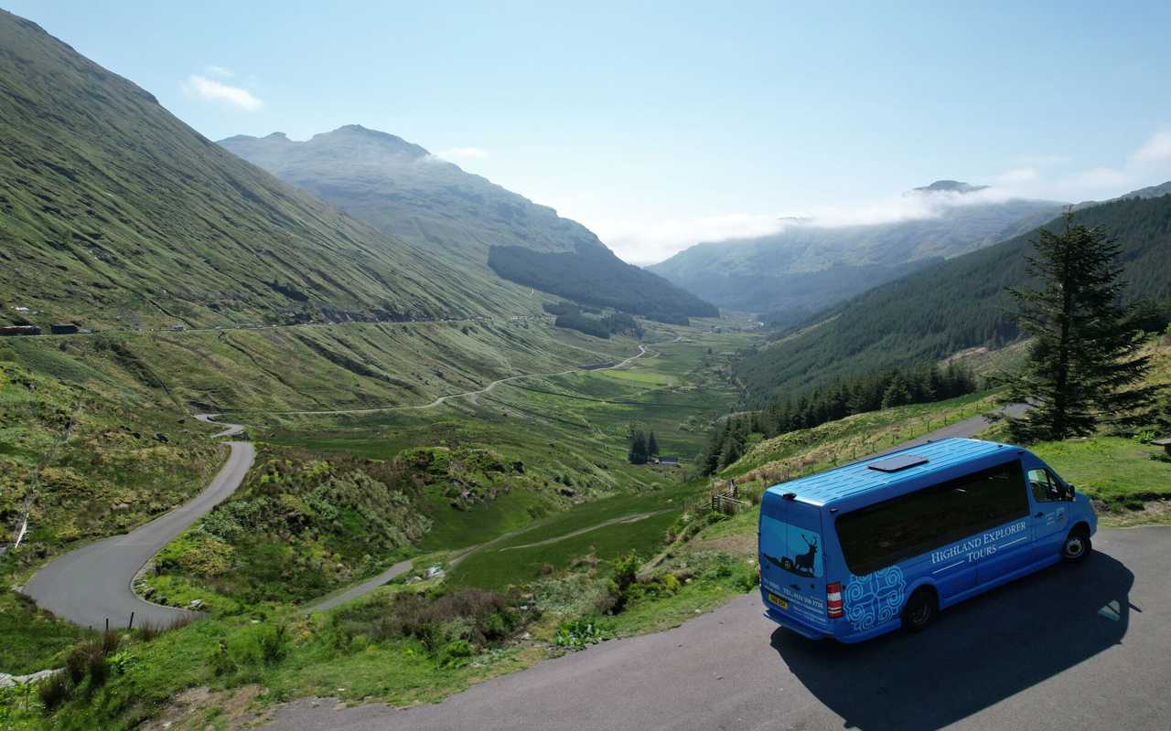 A blue Highland Explorer Tours van parked on a road overlooking a vast, green mountain valley with a winding road and distant peaks under a clear sky.